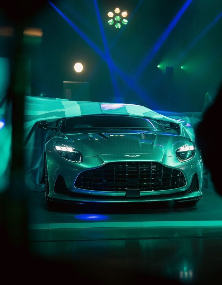 An Aston martin car which looks grey and matt green due to the room lighting. Image is taken from the front of the can with half the car uncovered